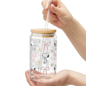 May include: A clear glass tumbler with a wooden lid and a glass straw. The tumbler is decorated with a repeating pattern of Snoopy characters, pink bows, and white flowers. The tumbler is held in a person's hands.