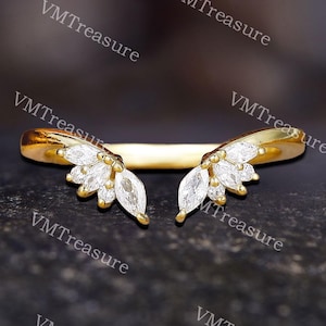 Op de afbeelding: Een goudkleurige ring met een gebogen ontwerp. De ring heeft twee clusters van witte edelstenen, elk lijkend op een gestileerde blad- of bloemblaadjesarrangement. De ring is geplaatst tegen een donkere, getextureerde achtergrond.