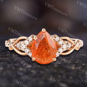 May include: A rose gold ring featuring a pear-shaped, orange gemstone center. The ring has a braided design on the band, accented with small, clear gemstones. The ring is set against a dark, textured background.