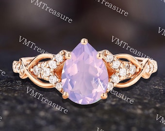 May include: Rose gold ring featuring a pear-shaped, light purple gemstone as the centerpiece. The ring is adorned with small, clear accent stones and intricate, twisted metalwork on the band. The ring is set against a dark, textured background.