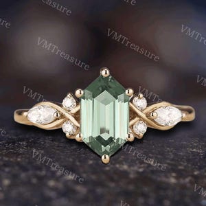 May include: A gold ring featuring a large, faceted, light green gemstone in a hexagonal shape. The ring is adorned with smaller white gemstones and decorative gold swirls on either side of the central stone. The ring is set against a dark, textured background.