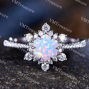 May include: A silver ring with a large, iridescent opal center stone, surrounded by smaller clear gemstones in a snowflake design. The band is encrusted with tiny clear stones. The ring is set against a dark, textured background.