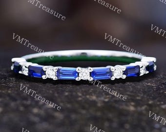 May include: A silver band ring featuring alternating blue baguette-cut gemstones and small round white gemstones. The ring is set against a dark, textured background, highlighting the sparkle of the stones.