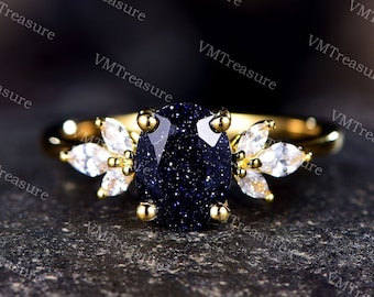 May include: A gold ring with a large oval blue gemstone and smaller clear gemstones on the band. The ring is set on a dark textured surface.