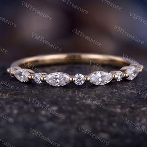 May include: A gold band ring featuring an alternating pattern of marquise-cut and round-cut clear stones. The stones are set along the top half of the thin metal band, creating a continuous, symmetrical design with a polished finish and clear sparkle.