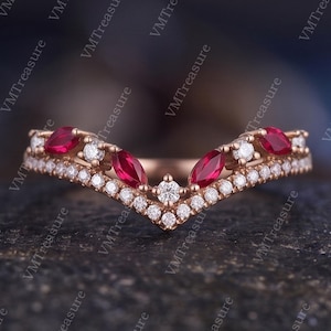 May include: A V-shaped ring crafted from rose-colored metal, featuring a row of small round white stones along the bottom edge and a series of alternating marquise-cut red stones and round white stones set along the top curved band.