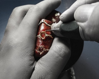 Può includere: Primo piano di una mano che tiene tre anelli con pietre preziose rosse e dettagli di diamanti. Gli anelli vengono lavorati con un piccolo strumento.