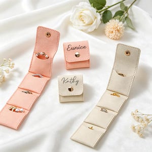 May include: Two foldable jewellery organisers in peach and cream colours, featuring gold-tone snap closures. The peach organiser is open, displaying rings, while the cream one is open with rings. Two closed pouches are labelled Eunice and Kathy.