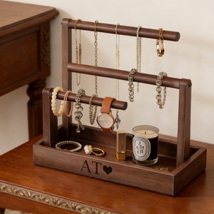 May include: A dark brown wooden jewelry organizer with multiple levels for necklaces, bracelets, and earrings. A watch, lipstick, and a candle are also displayed. The base of the organizer has the initials "AT" and a heart symbol.