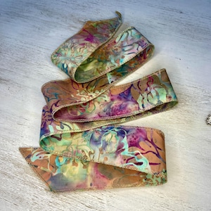 May include: A colorful ribbon with a batik-style pattern in shades of pink, purple, green, and brown. The edges are finished with a stitched trim. The ribbon is curled and layered, creating a decorative effect.