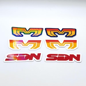 May include: Six retro-style stickers. The stickers feature the letter "M" in various colour gradients, including rainbow, orange, and yellow. Two stickers display the letters "SON" in red and pink.