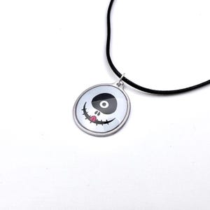 Pode incluir: Colar com pingente prateado com um desenho de rosto de desenho animado. O rosto tem um olho preto, um olho oval preto e uma boca sorridente com uma língua rosa. O pingente pende de um cordão preto. O fundo é branco.