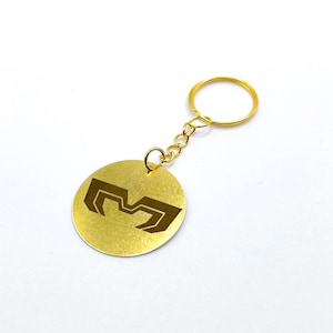 Peut inclure: Un porte-clés doré avec un pendentif circulaire. Le pendentif présente un motif géométrique marron foncé et est attaché à une chaîne et un anneau dorés. Le fond est blanc.