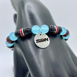May include: A bracelet with black, light blue, and maroon beads. The bracelet has a silver charm with the letters "SDN" and red and silver accents. The beads are strung on an elastic band.