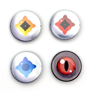 May include: Four round button pins with colorful designs. Three buttons feature a white background with a geometric shape in orange, red, and blue. The fourth button has a dark gray background with a red eye design.