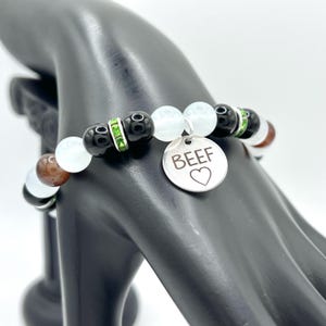 Puede incluir: Una pulsera de cuentas con cuentas negras, blancas y marrones, acentuada con espaciadores de pedrería verdes. Un dije plateado con la palabra "BEEF" y un corazón cuelga de la pulsera. La pulsera se muestra en una mano de maniquí negra.