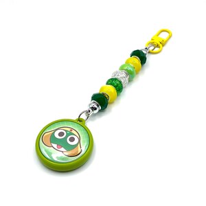 Può includere: Un portachiavi con perline verdi e gialle con un personaggio di rana dei cartoni animati su un ciondolo rotondo. Il ciondolo è verde con uno sfondo bianco e verde. Il portachiavi ha una chiusura gialla e una varietà di perline verdi, gialle e trasparenti.