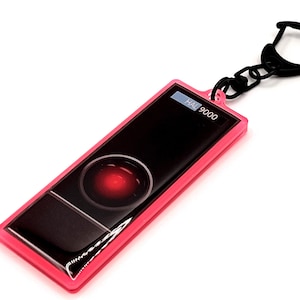 May include: A pink keychain with a black chain and a rectangular charm featuring a red light and the text "HAL 9000".