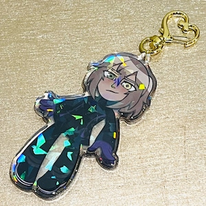 May include: A holographic charm featuring a cartoon character with grey hair, wearing a black and green outfit. The charm is attached to a gold heart-shaped keychain.