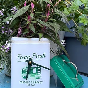 May include: White metal tin with a green tractor design and the text "Farm Fresh Produce & Market Est. 1779". The tin has a clock face with gold hands and a green lid.