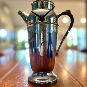 May include: A silver cocktail shaker with a clock face on the front. The clock hands are black and the numbers are red. The shaker has a spout and a handle.