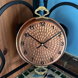 May include: A copper-colored wall clock with a hammered texture face and black hands. The clock has black numbers and gold-colored handles. The clock is mounted on a black metal frame, with a mosaic tile surface below.
