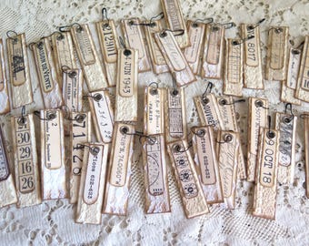 May include: A collection of vintage-style paper tags with various printed text and numbers. The tags are rectangular with torn edges, featuring black eyelets and attached black strings. The tags have a cream and beige color scheme.