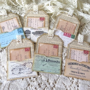 May include: A collection of vintage-style paper tags with various designs. Each tag features a small pocket holding a postcard or document. The tags are adorned with lace and text, creating a rustic aesthetic.