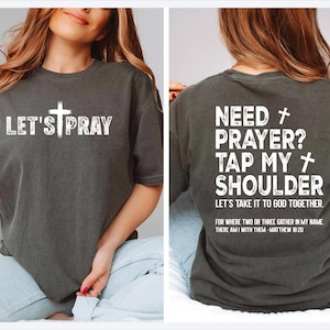 May include: Dark gray t-shirt with white text and a cross graphic. The front reads "LET'S PRAY". The back of the shirt says "NEED PRAYER? TAP MY SHOULDER" with a Bible verse. A casual, faith-based apparel item.