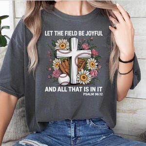 May include: Dark gray t-shirt with the text "LET THE FIELD BE JOYFUL AND ALL THAT IS IN IT" and Psalm 96:12. The design features a white cross with a baseball, glove, and flowers.