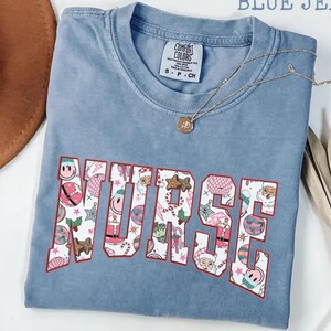 Könnte beinhalten: Ein blaues Comfort Colors T-Shirt mit dem Wort "NURSE" in einem festlichen Muster. Das Design enthält weihnachtliche Illustrationen in Rot, Weiß und Rosa. Eine goldene Halskette mit Anhänger liegt auf dem Shirt.