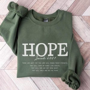 May include: Olive green sweatshirt with the word "HOPE" in large white letters. Below the word "HOPE" is the text "Isaiah 40:31" and a quote from the Bible. The sleeves are cuffed.