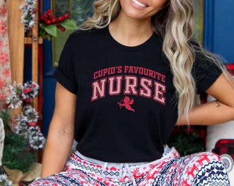 以下が含まれることがあります： 赤とピンクの文字で「Cupid's Favourite Nurse」と書かれた黒いTシャツ。テキストの下には、弓と矢を持ったキューピッドの小さなグラフィックがあります。このシャツは、柄物のパジャマパンツと組み合わせています。