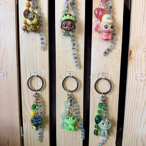 May include: Six metal keychains featuring small character figurines and beaded strands. The beads include lettered cubes spelling phrases like 'BIG EAVY VIBE', 'DREAMS COME TRUE', 'PINK PERFECTION', 'SHINE BRIGHT', 'KISSIN FROGS', and 'ROYAL BLISS' with various colored accents.