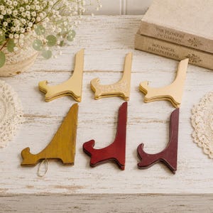 May include: A collection of six folding display stands arranged in two rows. The top row features three metallic gold stands, one with an embossed floral pattern. The bottom row includes one mustard yellow wooden stand and two dark red wooden stands.