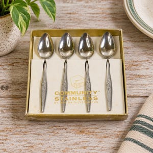 Peut inclure: Un ensemble de quatre cuillères en acier inoxydable argenté disposées dans une boîte dorée. La boîte porte l'inscription COMMUNITY STAINLESS BY ONEIDA SILVERSMITHS. Chaque manche de cuillère présente une finition texturée avec un motif de feuilles.