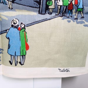 Puede incluir: Un pa&ntilde;o de cocina de lino blanco con una colorida ilustraci&oacute;n serigrafiada de personas caminando por una calle. El dise&ntilde;o incluye figuras con abrigos azules, verdes y rojos, con la palabra Ulster impresa en negro en la esquina inferior.
