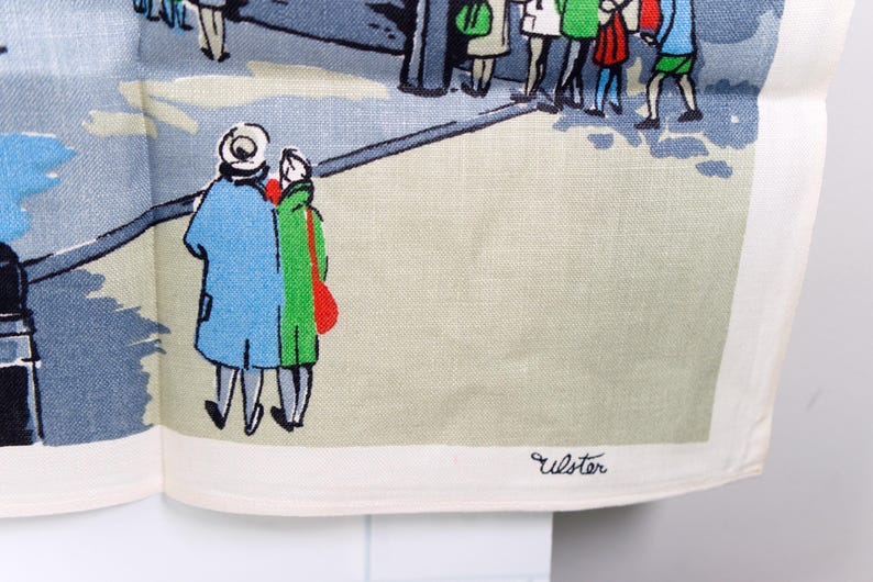 Puede incluir: Un pa&ntilde;o de cocina de lino blanco con una colorida ilustraci&oacute;n serigrafiada de personas caminando por una calle. El dise&ntilde;o incluye figuras con abrigos azules, verdes y rojos, con la palabra Ulster impresa en negro en la esquina inferior.