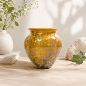 May include: A glazed ceramic vase with a rounded body and a narrow neck. The vase has a mottled pattern in gold, brown, and green. It is displayed on a wooden surface. A small white vase and flowers are in the background.
