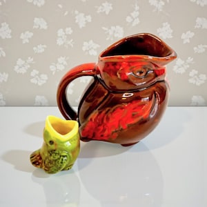 May include: A brown ceramic owl-shaped pitcher with a red accent and a matching small owl-shaped creamer. The pitcher has a handle and a spout. The creamer is green and yellow.