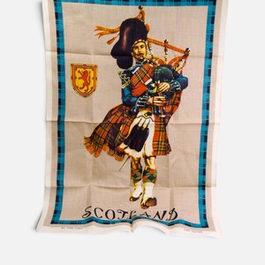Puede incluir: Un pa&ntilde;o de cocina rectangular con una ilustraci&oacute;n central de un gaitero con vestimenta tradicional escocesa, incluyendo kilt y gorro de piel de oso. El dise&ntilde;o incluye un emblema de le&oacute;n rojo, un borde de cuadros azul y negro y la palabra SCOTLAND.