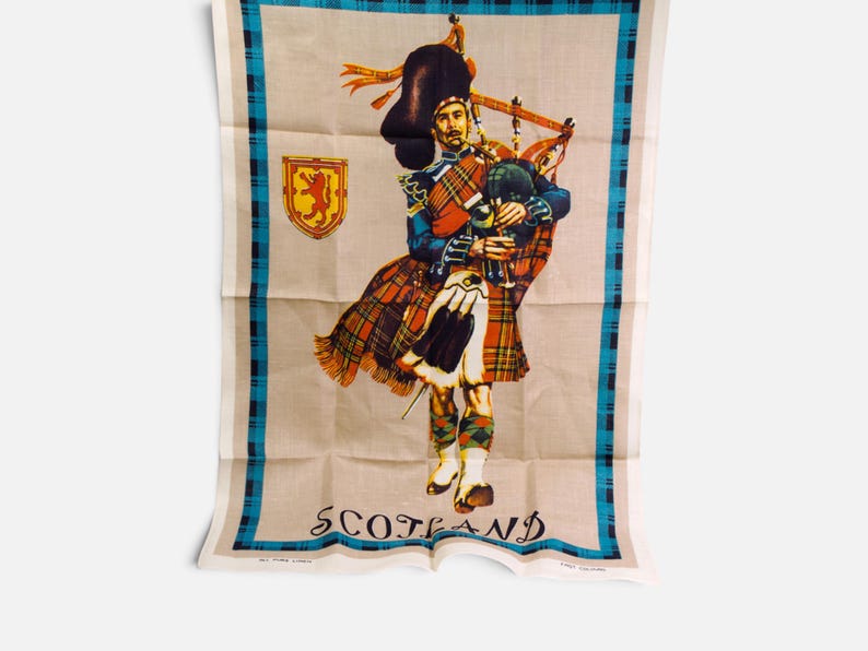 Puede incluir: Un pa&ntilde;o de cocina rectangular con una ilustraci&oacute;n central de un gaitero con vestimenta tradicional escocesa, incluyendo kilt y gorro de piel de oso. El dise&ntilde;o incluye un emblema de le&oacute;n rojo, un borde de cuadros azul y negro y la palabra SCOTLAND.