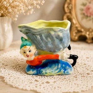 May include: A vintage ceramic planter in the shape of a whimsical figure. The planter features a blue and yellow container held by a small figure in a red shirt, blue trousers, and a green hat. The planter is painted with bright colours and has a glossy finish.