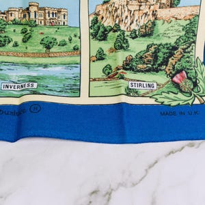 Puede incluir: Un textil con borde azul que presenta ilustraciones impresas de monumentos escoceses. El panel izquierdo muestra un castillo etiquetado como Inverness, y el derecho muestra un castillo etiquetado como Stirling con un cardo. Texto: Dunluce y Made in U.K.
