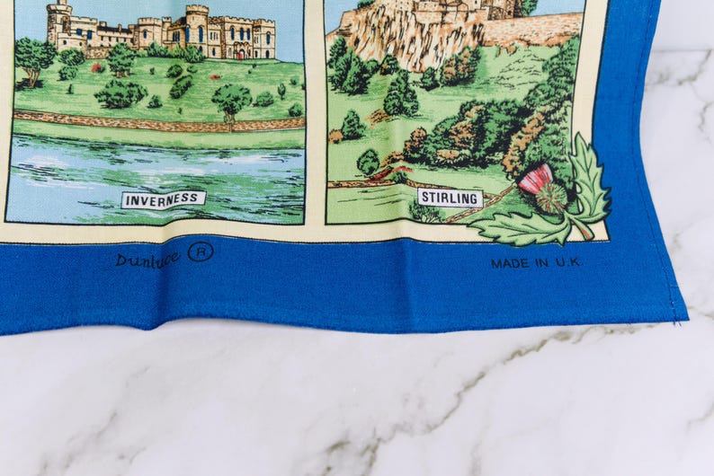 Puede incluir: Un textil con borde azul que presenta ilustraciones impresas de monumentos escoceses. El panel izquierdo muestra un castillo etiquetado como Inverness, y el derecho muestra un castillo etiquetado como Stirling con un cardo. Texto: Dunluce y Made in U.K.