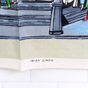 Puede incluir: Un pa&ntilde;o de cocina de lino irland&eacute;s con una colorida ilustraci&oacute;n gr&aacute;fica de personas caminando por una calle con farolas. La tela tiene un borde color crema claro en la parte inferior con el texto IRISH LINEN impreso en letras may&uacute;sculas negras.