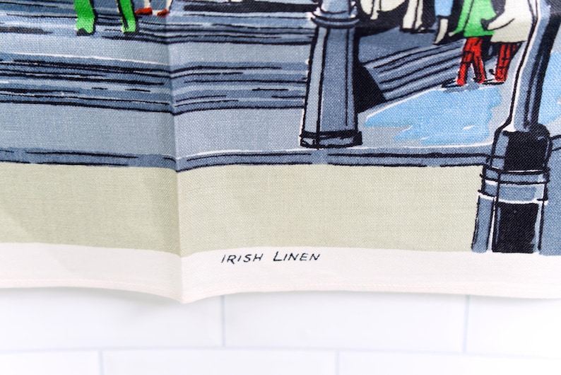 Puede incluir: Un pa&ntilde;o de cocina de lino irland&eacute;s con una colorida ilustraci&oacute;n gr&aacute;fica de personas caminando por una calle con farolas. La tela tiene un borde color crema claro en la parte inferior con el texto IRISH LINEN impreso en letras may&uacute;sculas negras.