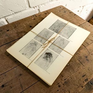 May include: A stack of off-white paper sheets tied together with thin brown twine. The top sheet features five distinct black and white sketches of figures in various poses, each accompanied by small printed text labels near the bottom corners.