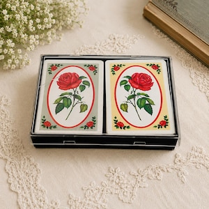 May include: A double deck of playing cards housed in a clear plastic case. Each deck features a central illustration of a red rose with green leaves enclosed in an oval frame, with one deck on a light gray background and the other on a cream background.