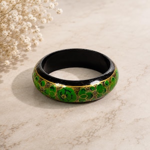 May include: A circular bangle bracelet with a glossy black base, featuring hand-painted vibrant green floral motifs and intricate gold line detailing throughout the exterior surface, showcasing a smooth, rounded profile and a solid black interior.